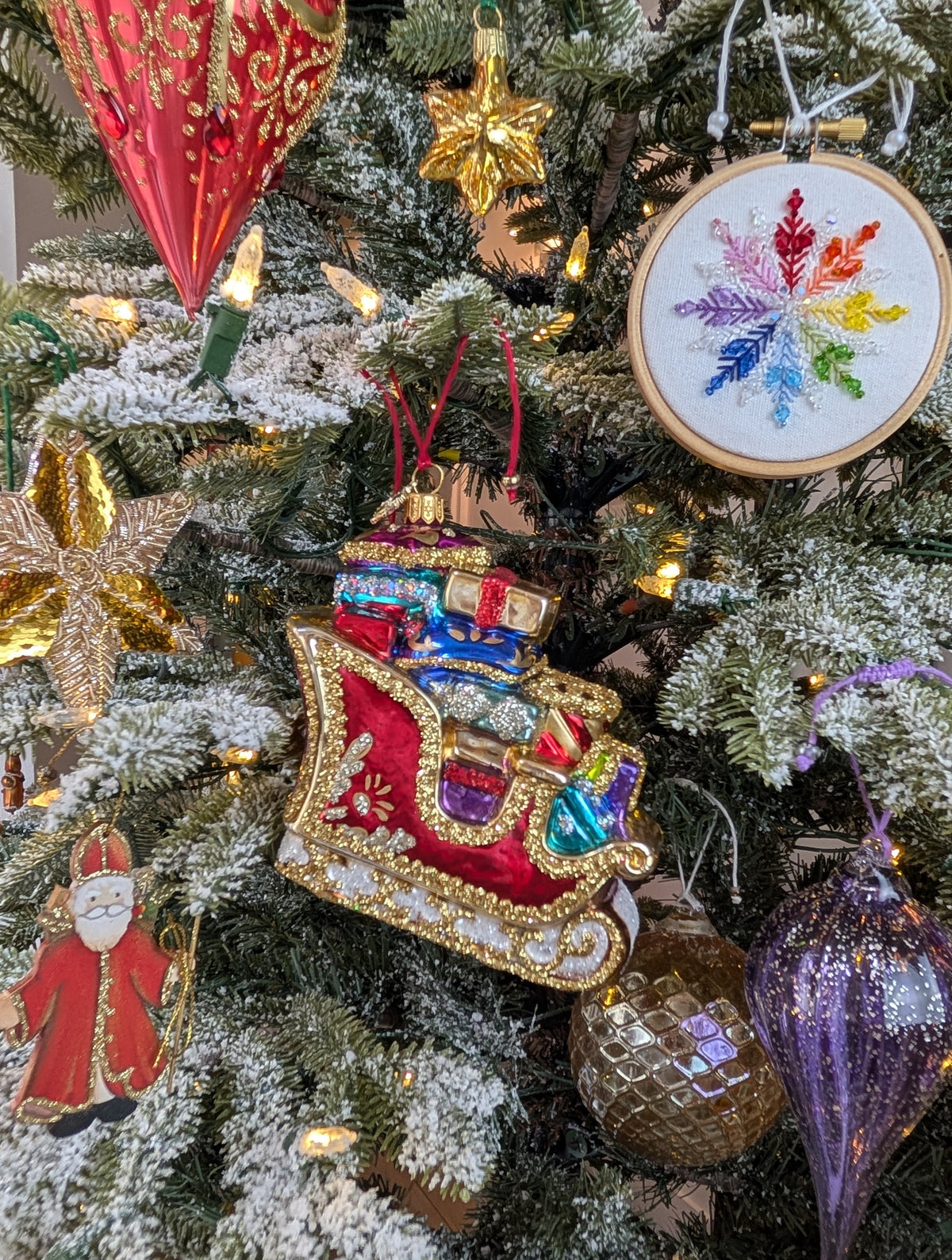 Suspension en forme de traineau du père Noël rouge et doré, garni de paquets cadeaux multicolores. Suspension brodée flocon arc-en-ciel en fil et cristaux. suspension miniature étoile dorée. Suspension en bois Saint Nicolas. Suspension en verre en forme de goutte violette pailletée argent et boule dorée style ananas.