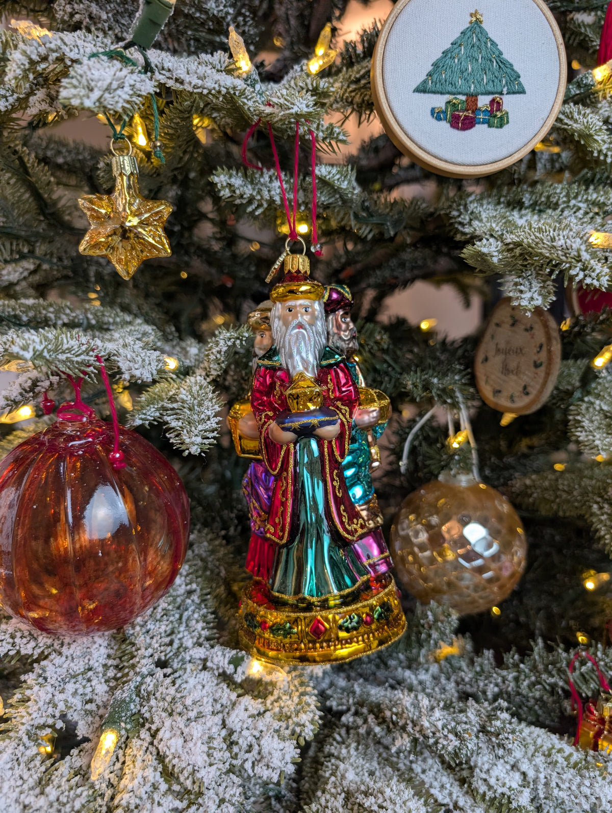 Suspension en forme des trois rois mages apportant leurs cadeaux. ils sont habillés de riches couleurs et chapeaux et sont posés sur un socle doré décoré de houx et baies rouges. Suspensions boules de noël en verre style venise rouge et style ananas or. Suspension miniature en forme d'étoile dorée. Suspension broderie sapin décoré de bougies, étoile dorée au sommet et paquets cadeaux au pied du sapin. Suspension rondin de bois pyrogravé joyeux noël.