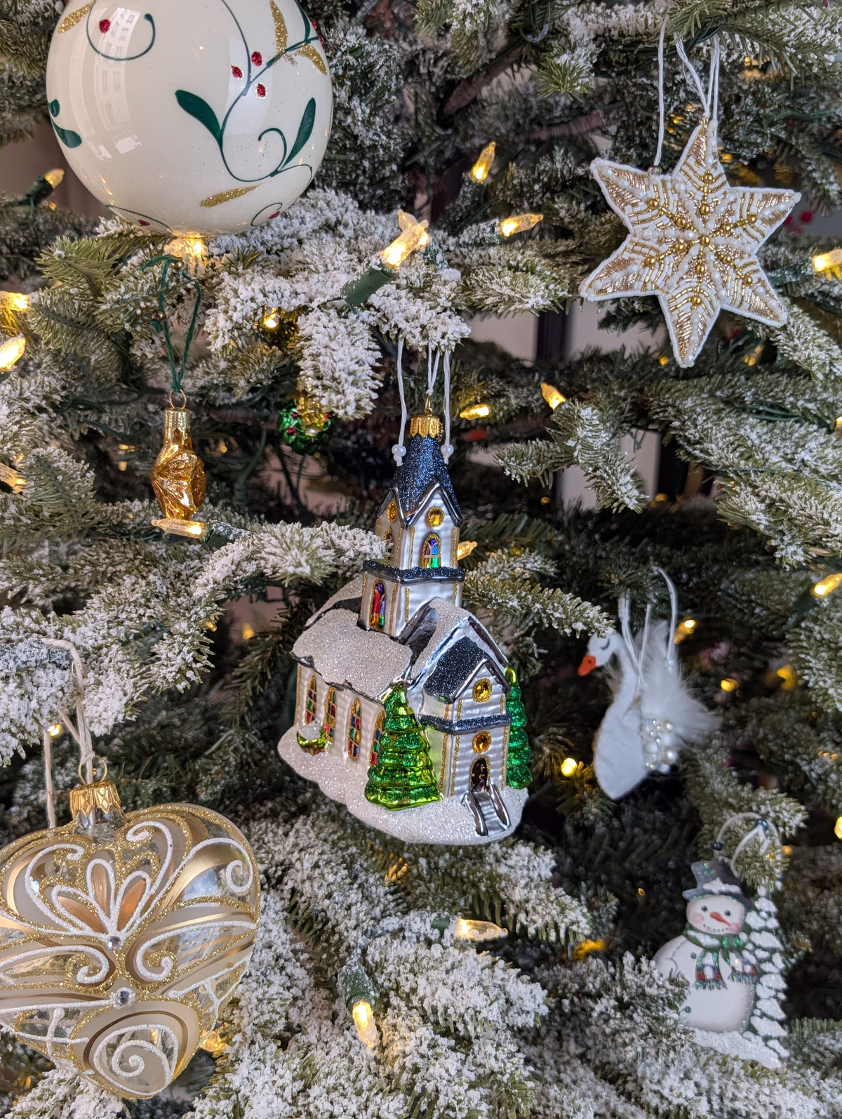 Suspension en forme d'église blanche, avec des vitraux colorés vert, jaune, bleu, violet et rouge et des rosaces dorée, son toit bleu gris est enneigé et l'église est entourée de sapin verts; étoile brodée de perles blanches et or ; cygne en feutrine avec des plumes blanches serties de perles et cristaux ; miniature en forme d'étoile dorée; boule de Noël crème décorée de feuilles de gui vertes et or avec des baies rouges ; cœur transparent volutes baroques or blanc et argent ; bonhomme de neige et son sapin