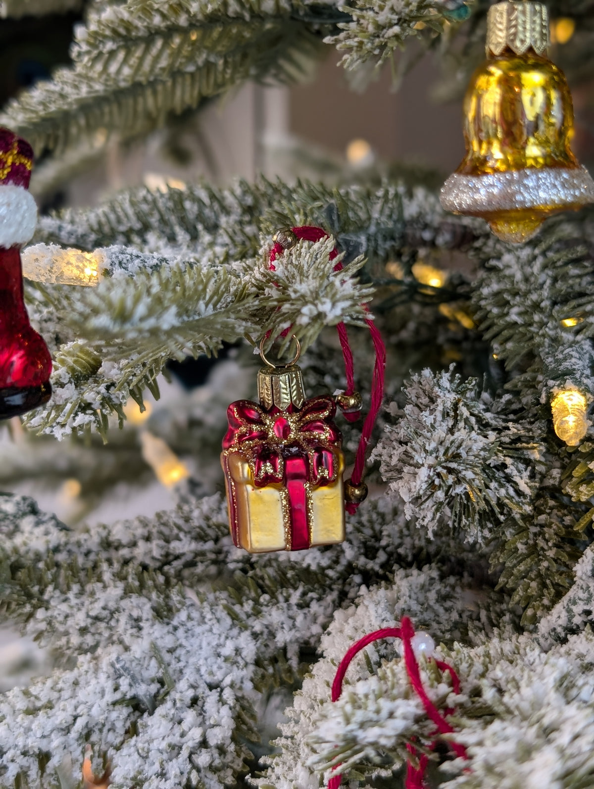 Suspension de Noël en verre soufflé, chromée et peinte à la main en forme de paquet cadeau doré avec un ruban rouge aux liserés dorés pailletés qui se termine par un nœud sur le dessus du cadeau. Suspension de Noël en verre soufflé, chromée et peinte à la main en forme de cloche dorée et paillettes argentées.