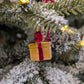 Suspension de Noël en verre soufflé, chromée et peinte à la main en forme de paquet cadeau doré avec un ruban rouge aux liserés dorés pailletés qui se termine par un nœud sur le dessus du cadeau. 