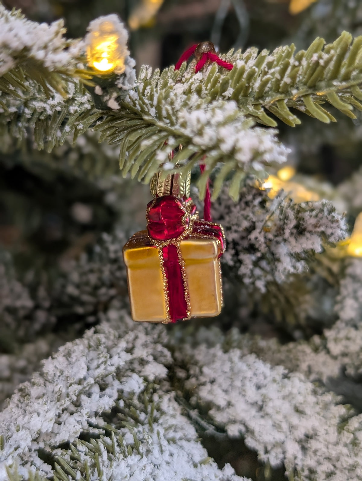 Suspension de Noël en verre soufflé, chromée et peinte à la main en forme de paquet cadeau doré avec un ruban rouge aux liserés dorés pailletés qui se termine par un nœud sur le dessus du cadeau. 