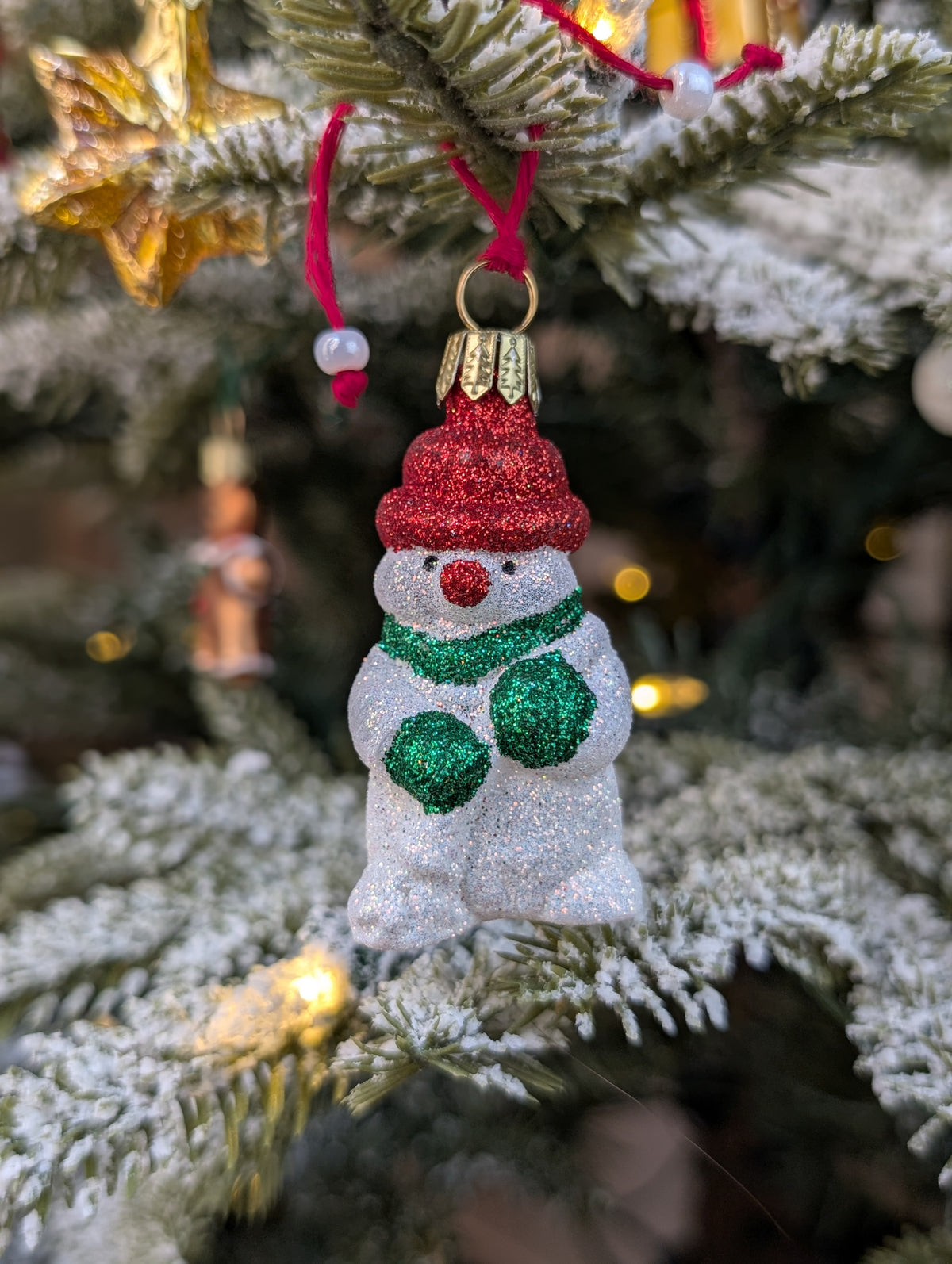 Suspension de Noël en verre soufflé puis pailleté en forme de bonhomme de neige avec un bonnet et nez rouges et une écharpe et des gants verts. 