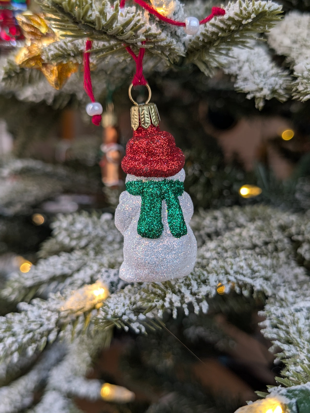 Dos de la suspension de Noël en verre soufflé puis pailleté en forme de bonhomme de neige avec un bonnet rouge et une écharpe verte nouée dans le dos. 