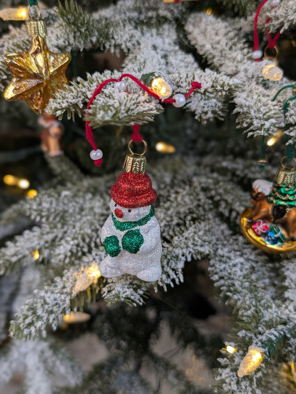 Vu de hauteur de la suspension de Noël en verre soufflé puis pailleté en forme de bonhomme de neige avec un bonnet et nez rouges et une écharpe et des gants verts. 