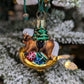 Suspension en verre soufflé, chromée et peinte à la main en forme de cheval à bascule avec une crinière blanche et une selle noire. Il a des paquets cadeaux entre les pattes et un sapin décoré d'une guirlande dorée sur le dos. 