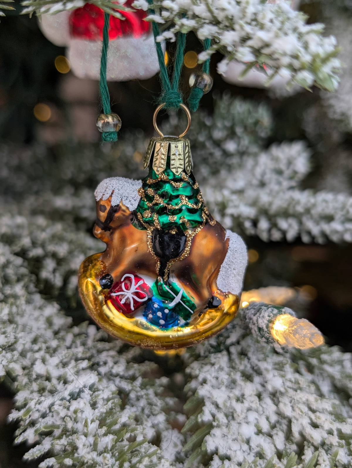 Suspension en verre soufflé, chromée et peinte à la main en forme de cheval à bascule avec une crinière blanche et une selle noire. Il a des paquets cadeaux entre les pattes et un sapin décoré d'une guirlande dorée sur le dos. 