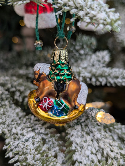 Suspension en verre soufflé, chromée et peinte à la main en forme de cheval à bascule avec une crinière blanche et une selle noire. Il a des paquets cadeaux entre les pattes et un sapin décoré d'une guirlande dorée sur le dos. 