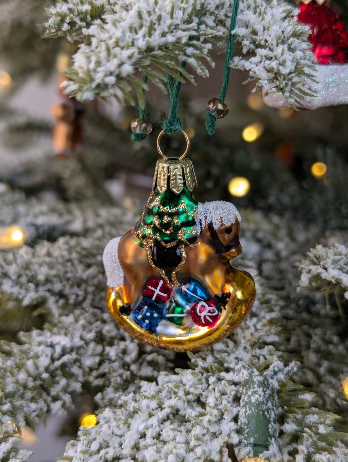 Suspension en verre soufflé, chromée et peinte à la main en forme de cheval à bascule avec une crinière blanche et une selle noire. Il a des paquets cadeaux entre les pattes et un sapin décoré d'une guirlande dorée sur le dos. 