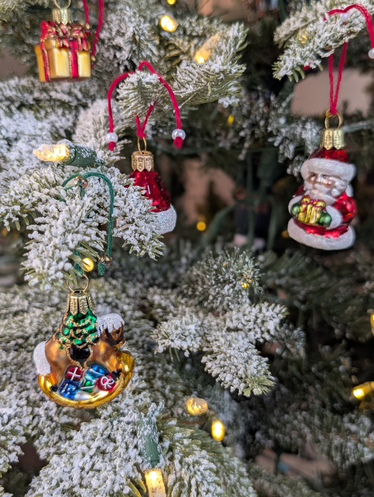 Suspension en verre soufflé, chromée et peinte à la main en forme de cheval à bascule avec une crinière blanche et une selle noire. Il a des paquets cadeaux entre les pattes et un sapin décoré d'une guirlande dorée sur le dos. Suspensions en verre soufflé, chromée et peintes à la main en forme de père Noël et de paquet cadeaux doré serti d'un ruban et nœud rouge.