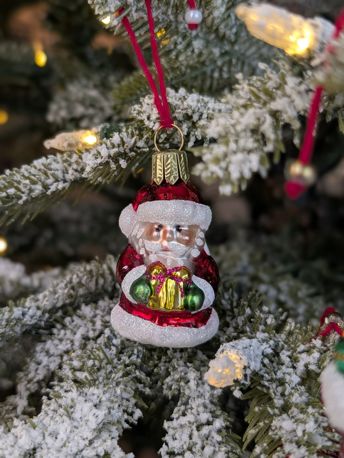 Suspension en verre soufflé, chromée et peinte à la main en forme de père Noël vêtu de son habit rouge, de gants verts et portant un cadeau doré serti d'un nœud rouge. La fourrure de son habit et de son bonnet est représenté en paillettes blanches.
