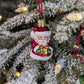 Suspension en verre soufflé, chromée et peinte à la main en forme de père Noël vêtu de son habit rouge, de gants verts et portant un cadeau doré serti d'un nœud rouge. La fourrure de son habit et de son bonnet est représenté en paillettes blanches.