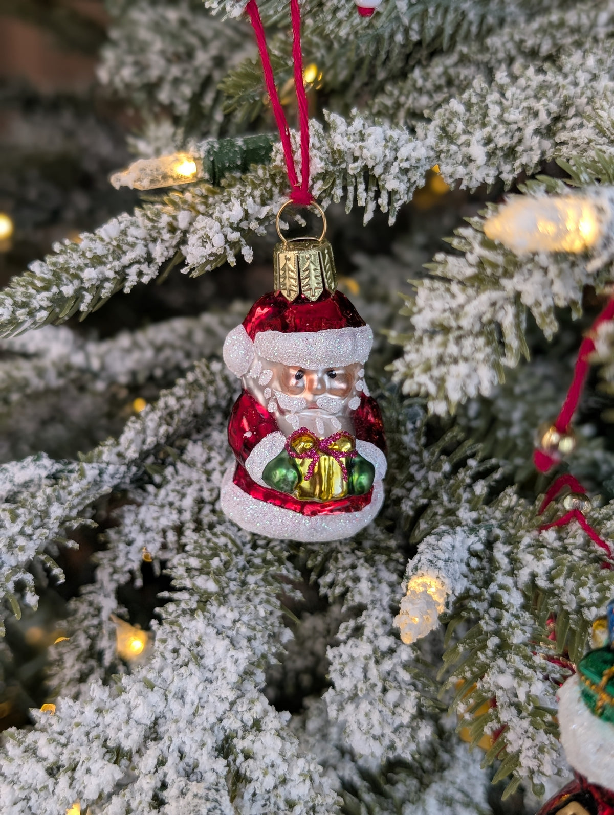 Suspension en verre soufflé, chromée et peinte à la main en forme de père Noël vêtu de son habit rouge, de gants verts et portant un cadeau doré serti d'un nœud rouge. La fourrure de son habit et de son bonnet est représenté en paillettes blanches.