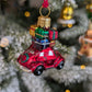 Suspension de Noël en verre soufflé, chromée et peinte à la main en forme de voiture rouge avec des paquets cadeaux rouge, or, bleu et vert sur son toit et une couronne de Noël accrochée à l'arrière sur le coffre.