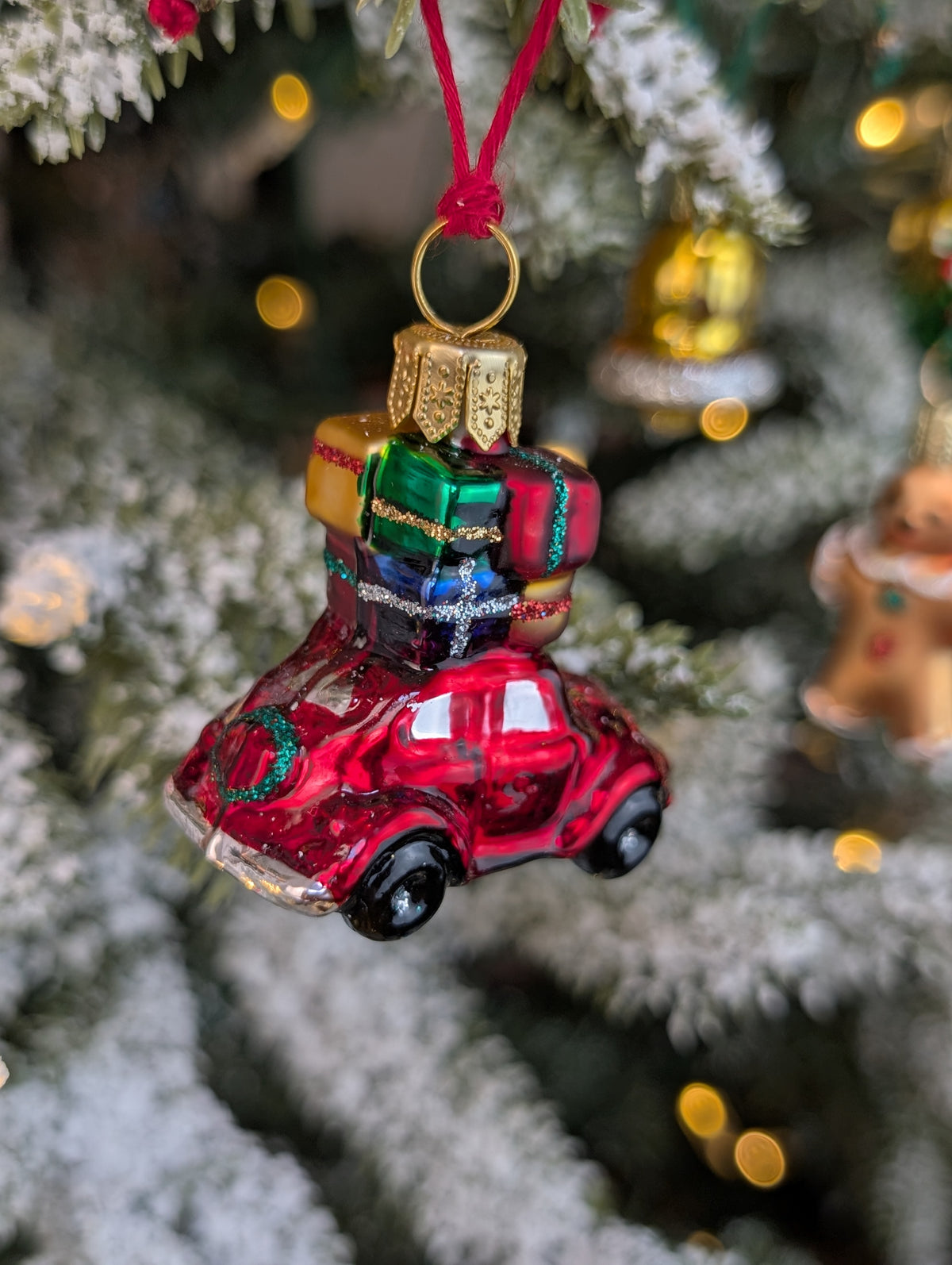 Suspension de Noël en verre soufflé, chromée et peinte à la main en forme de voiture rouge avec des paquets cadeaux rouge, or, bleu et vert sur son toit et une couronne de Noël accrochée à l'arrière sur le coffre.