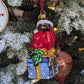 Suspension en forme d'oiseau cardinal rouge qui porte un bonnet de noël avec fourrure et pompon blanc, assis sur un cadeau bleu décoré de flocons blancs et sertie d'un nœud doré et de feuilles de houx.