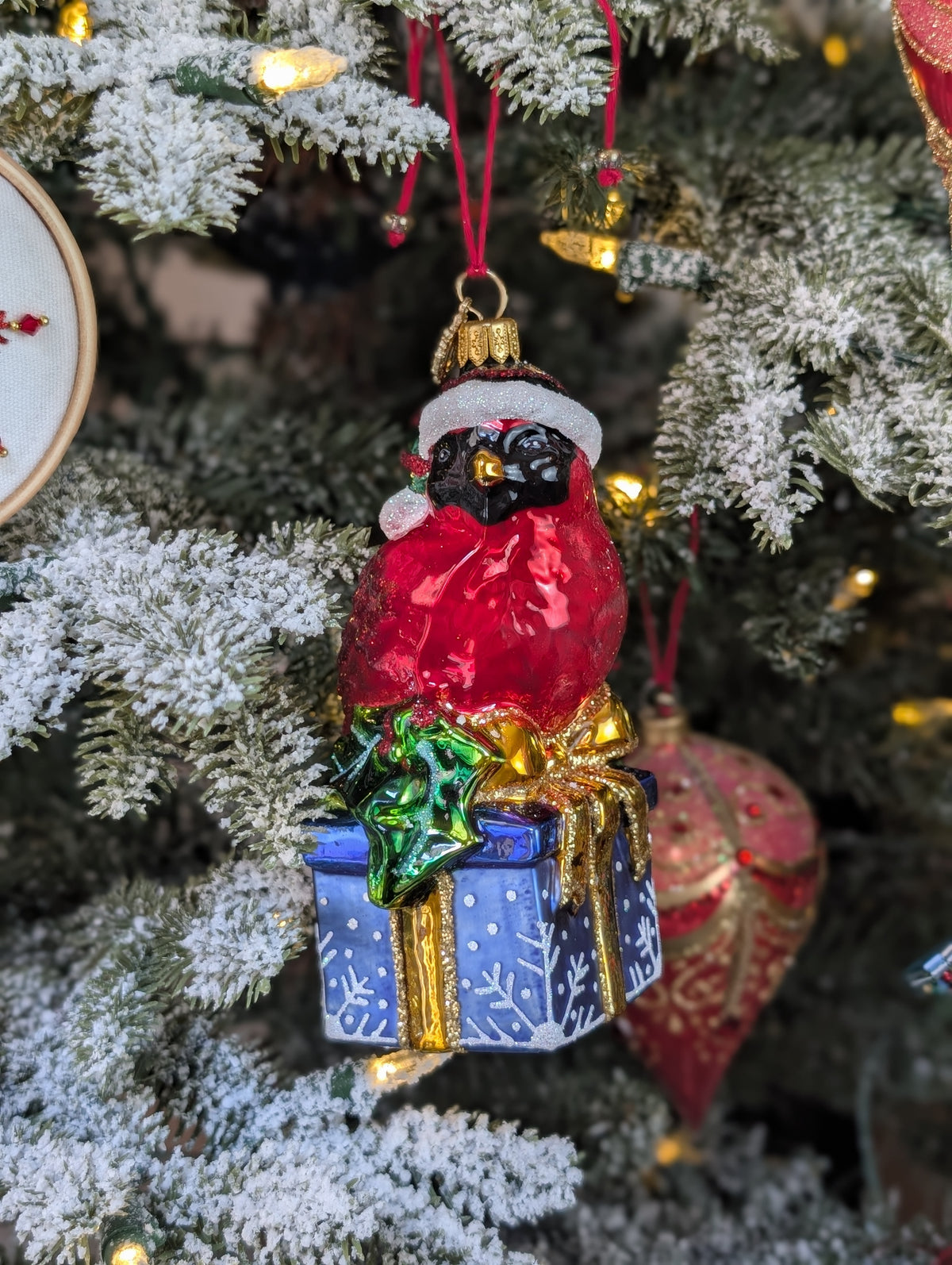 Suspension en forme d'oiseau cardinal rouge qui porte un bonnet de noël avec fourrure et pompon blanc, assis sur un cadeau bleu décoré de flocons blancs et sertie d'un nœud doré et de feuilles de houx.