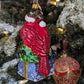 Dos de la suspension en forme d'oiseau cardinal rouge qui porte un bonnet de noël vert rayé de rouge avec fourrure et pompon blanc, assis sur un cadeau bleu décoré de flocons blancs et sertie d'un nœud doré et de feuilles de houx.
