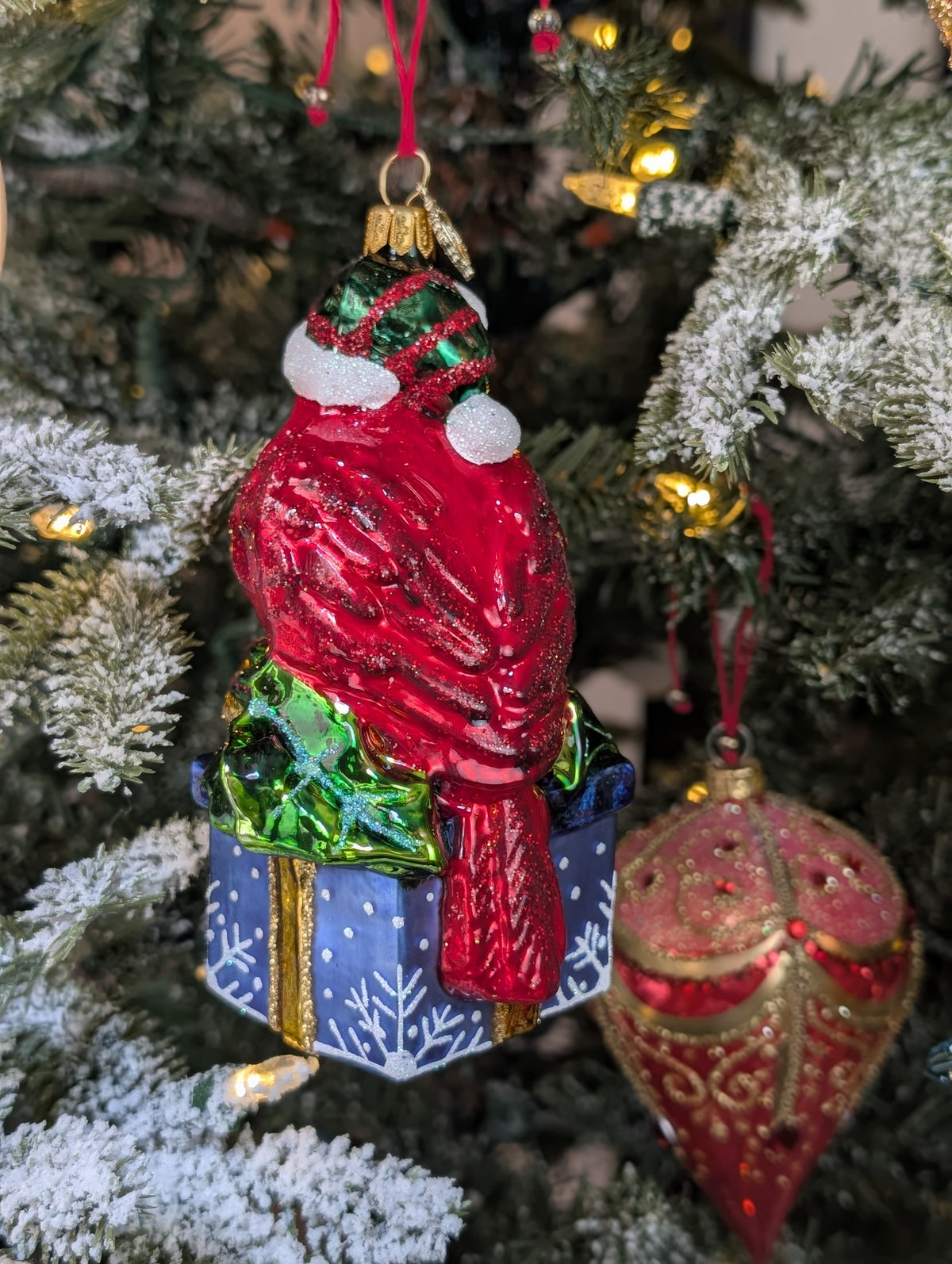 Dos de la suspension en forme d'oiseau cardinal rouge qui porte un bonnet de noël vert rayé de rouge avec fourrure et pompon blanc, assis sur un cadeau bleu décoré de flocons blancs et sertie d'un nœud doré et de feuilles de houx.