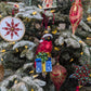 Suspension en forme d'oiseau cardinal rouge qui porte un bonnet de noël vert rayé de rouge avec fourrure et pompon blanc, assis sur un cadeau bleu décoré de flocons blancs et sertie d'un nœud doré et de feuilles de houx. Suspension brodée flocon rouge et or, fil métallisé, perles et cristaux. Suspensions en forme de gouttes rouge décorée de paillettes or dans un style baroque. Suspension en forme d'étoile brodée de perles vertes et rouges et de cristaux rouge, finie par une pampille rouge