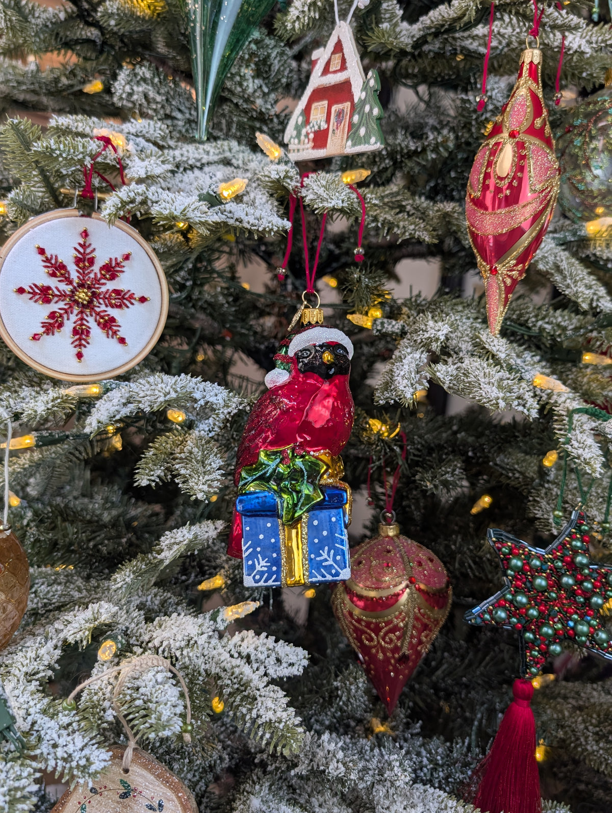 Suspension en forme d'oiseau cardinal rouge qui porte un bonnet de noël vert rayé de rouge avec fourrure et pompon blanc, assis sur un cadeau bleu décoré de flocons blancs et sertie d'un nœud doré et de feuilles de houx. Suspension brodée flocon rouge et or, fil métallisé, perles et cristaux. Suspensions en forme de gouttes rouge décorée de paillettes or dans un style baroque. Suspension en forme d'étoile brodée de perles vertes et rouges et de cristaux rouge, finie par une pampille rouge