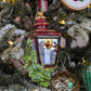 Suspension en forme de lanterne rouge et or avec une bougie allumée à l'intérieur et des branches de sapin et baies rouges sur les cotés. 