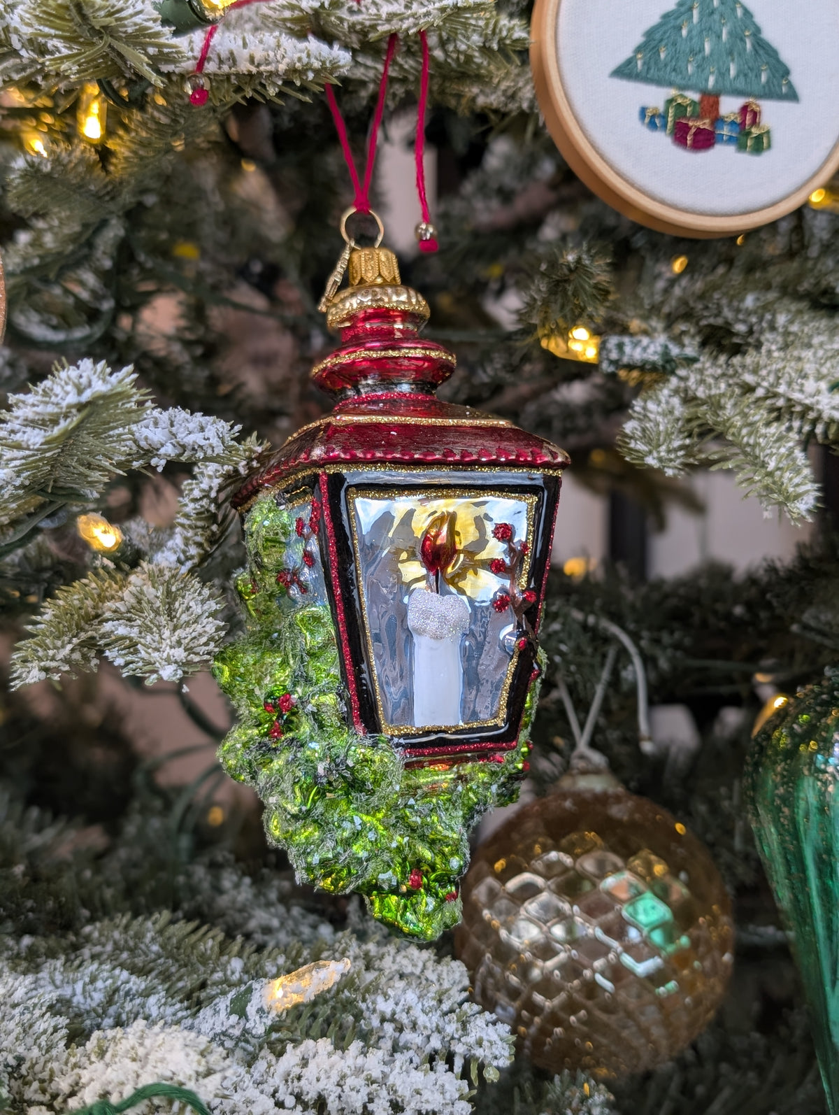 Suspension en forme de lanterne rouge et or avec une bougie allumée à l'intérieur et des branches de sapin et baies rouges sur les cotés. 