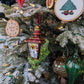 Suspensions en forme de lanterne rouge et or avec une bougie allumée à l'intérieur et des branches de sapin et baies rouges sur les cotés ; rondin de bois gravé Joyeux Noël avec couronne de noël autour ; rondin de bois avec un hérisson qui tient un cadeau et regarde un cœur rouge ; broderie d'un sapin de noël décoré de bougies blanches allumées et d'une étoile en son sommet, des cadeaux bordeaux, bleus et verts sont à son pied; goutte en verre verte pailletée or; boule en verre or style ananas.