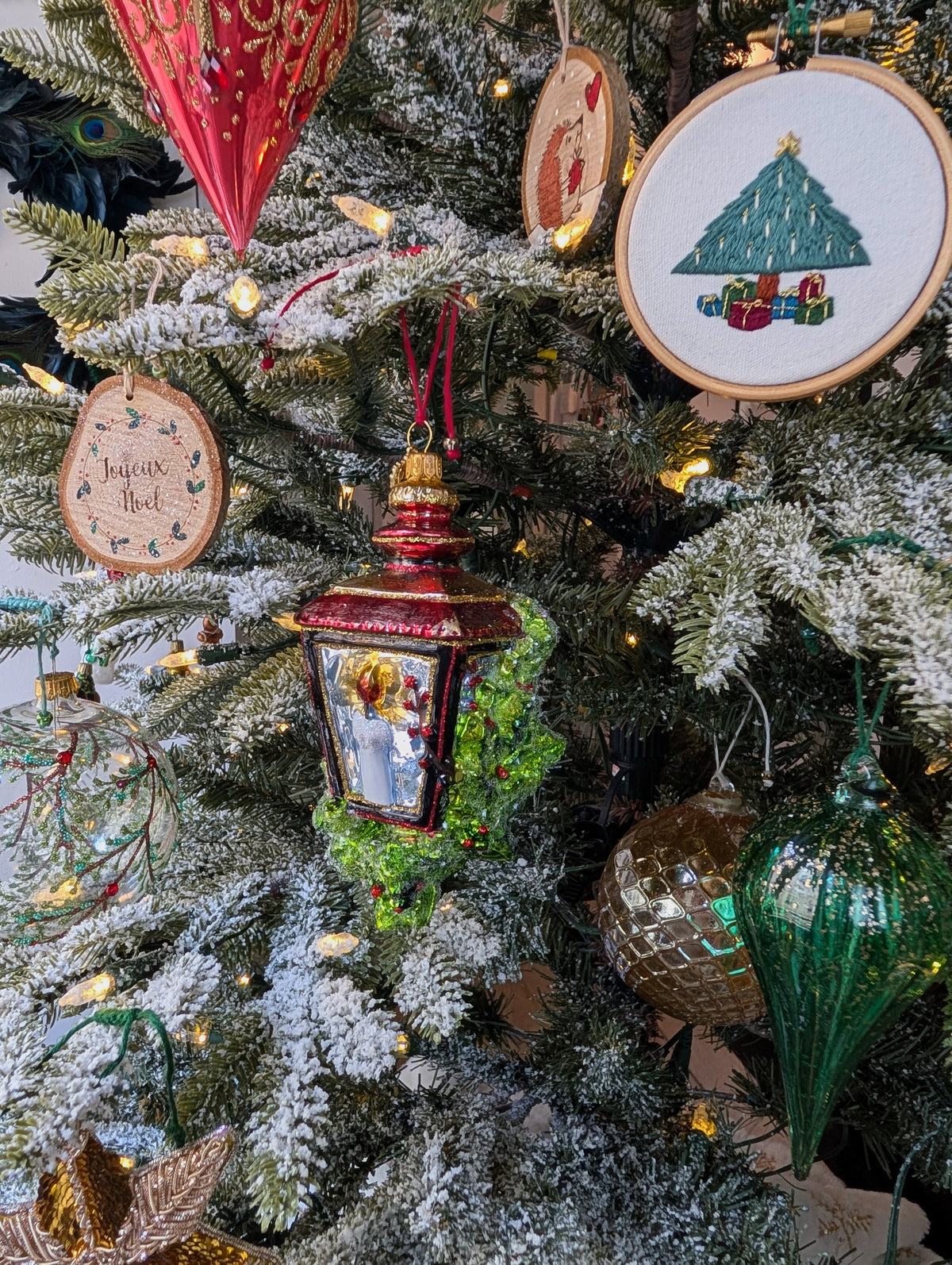 Suspensions en forme de lanterne rouge et or avec une bougie allumée à l'intérieur et des branches de sapin et baies rouges sur les cotés ; rondin de bois gravé Joyeux Noël avec couronne de noël autour ; rondin de bois avec un hérisson qui tient un cadeau et regarde un cœur rouge ; broderie d'un sapin de noël décoré de bougies blanches allumées et d'une étoile en son sommet, des cadeaux bordeaux, bleus et verts sont à son pied; goutte en verre verte pailletée or; boule en verre or style ananas.