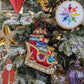 Suspension en forme de traineau du père Noël rouge et doré, garni de paquets cadeaux multicolores.  Suspension brodée flocon arc-en-ciel en fil et cristaux. suspension miniature étoile dorée. Suspension en bois Saint Nicolas. Suspension en verre en forme de goutte violette pailletée argent et boule dorée style ananas.