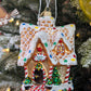 Suspension en forme de maison en pain d'épices décorée de glaçage blanc et de bonbons rouges, verts, bleus et jaunes. Joyeux Noël est écrit sur une pancarte au-dessus de la porte. 