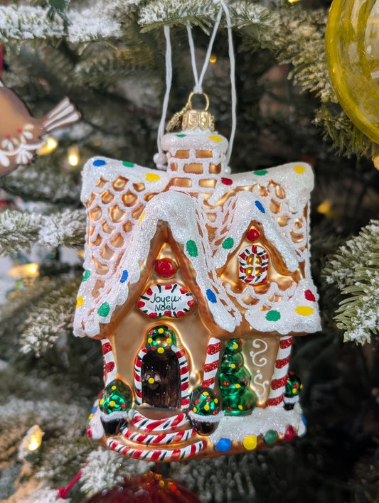 Suspension en forme de maison en pain d'épices décorée de glaçage blanc et de bonbons rouges, verts, bleus et jaunes. Joyeux Noël est écrit sur une pancarte au-dessus de la porte. 