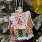 Suspension en forme de maison en pain d'épices décorée de glaçage blanc et de bonbons rouges, verts, bleus et jaunes. Joyeux Noël est écrit sur une pancarte au-dessus de la porte. 