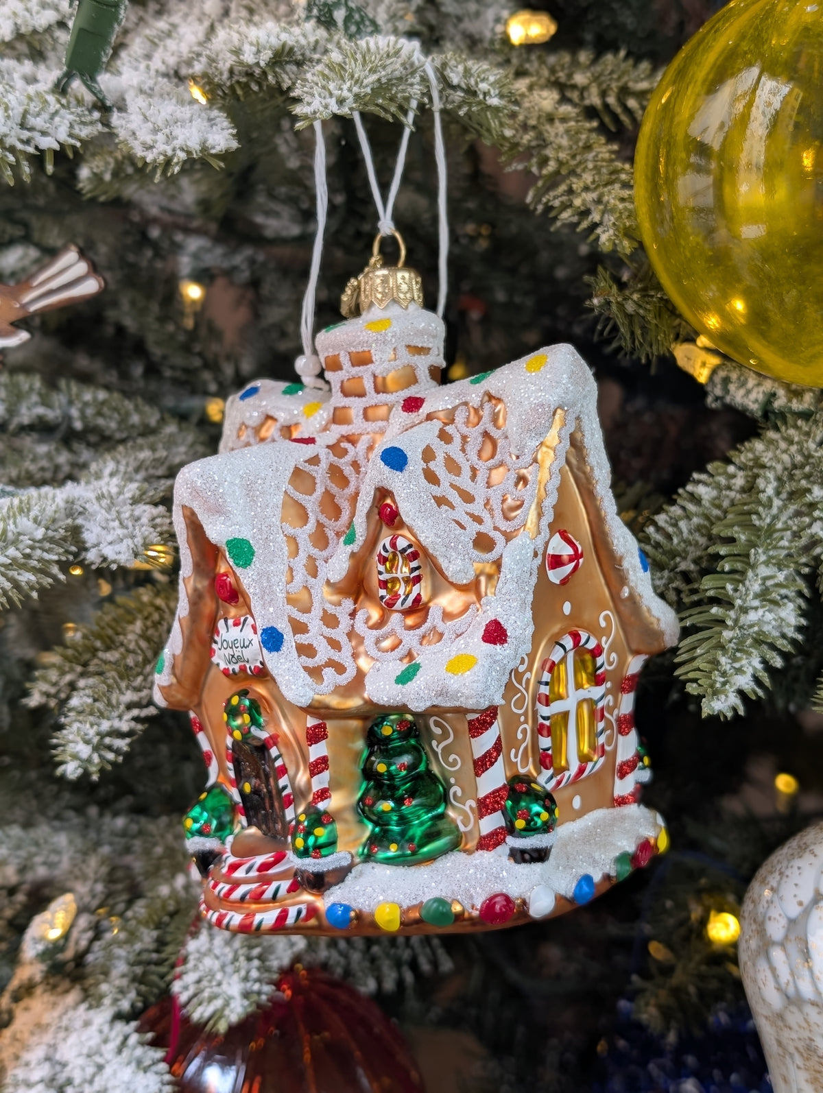 Suspension en forme de maison en pain d'épices décorée de glaçage blanc et de bonbons rouges, verts, bleus et jaunes. Joyeux Noël est écrit sur une pancarte au-dessus de la porte. 