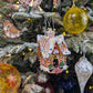 Suspension en forme de maison en pain d'épices décorée de glaçage blanc et de bonbons rouges, verts, bleus et jaunes. Joyeux Noël est écrit sur une pancarte au-dessus de la porte. Cinq boules en verre : blanche et rouge, jaunes style venise, jaune et orange style ananas, bleu foncé style ananas; rouge style venise. Une suspension goutte en verre blanche et transparente avec paillettes dorées. Une suspension en bois en forme d'oiseau en pain d'épices. 