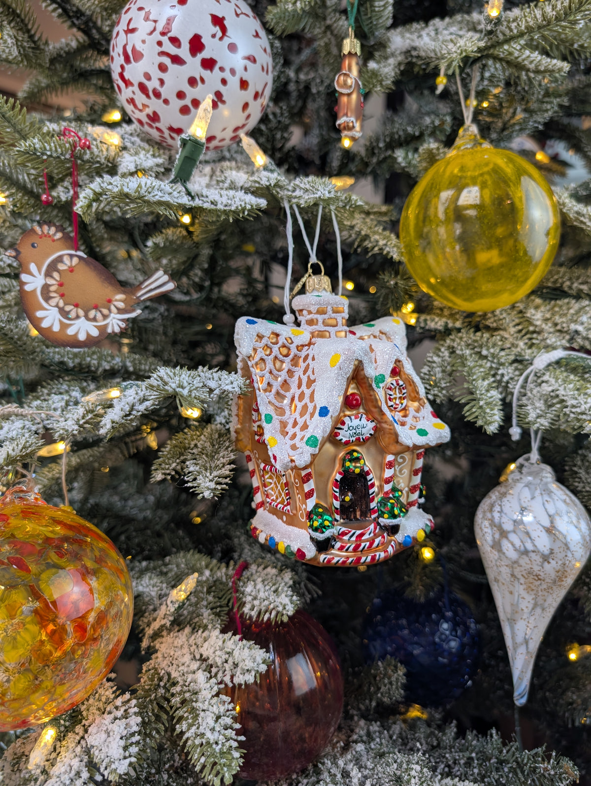 Suspension en forme de maison en pain d'épices décorée de glaçage blanc et de bonbons rouges, verts, bleus et jaunes. Joyeux Noël est écrit sur une pancarte au-dessus de la porte. Cinq boules en verre : blanche et rouge, jaunes style venise, jaune et orange style ananas, bleu foncé style ananas; rouge style venise. Une suspension goutte en verre blanche et transparente avec paillettes dorées. Une suspension en bois en forme d'oiseau en pain d'épices. 