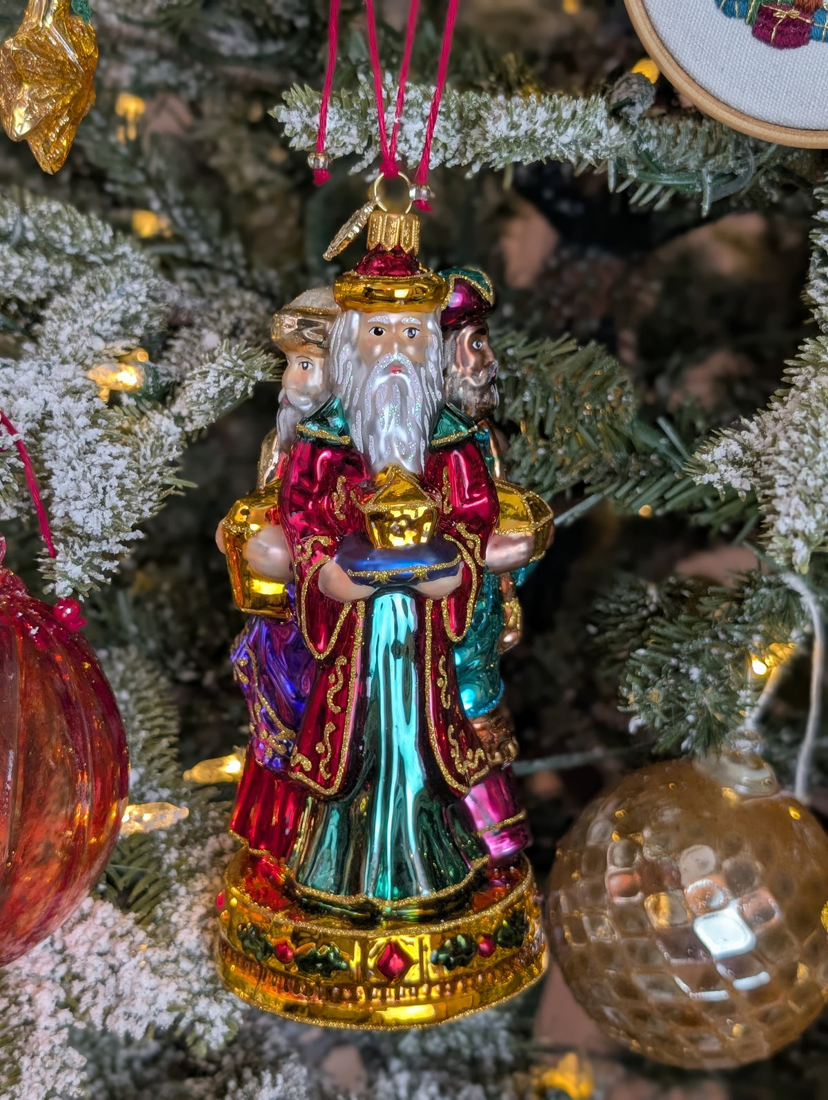 Suspension en forme des trois rois mages apportant leurs cadeaux. ils sont habillés de riches couleurs et chapeaux et sont posés sur un socle doré décoré de houx et baies rouges. Vue sur Melchior de face apportant l'or et Gaspard de profil apportant l'encens et Balthazar de profil offrant la myrrhe.