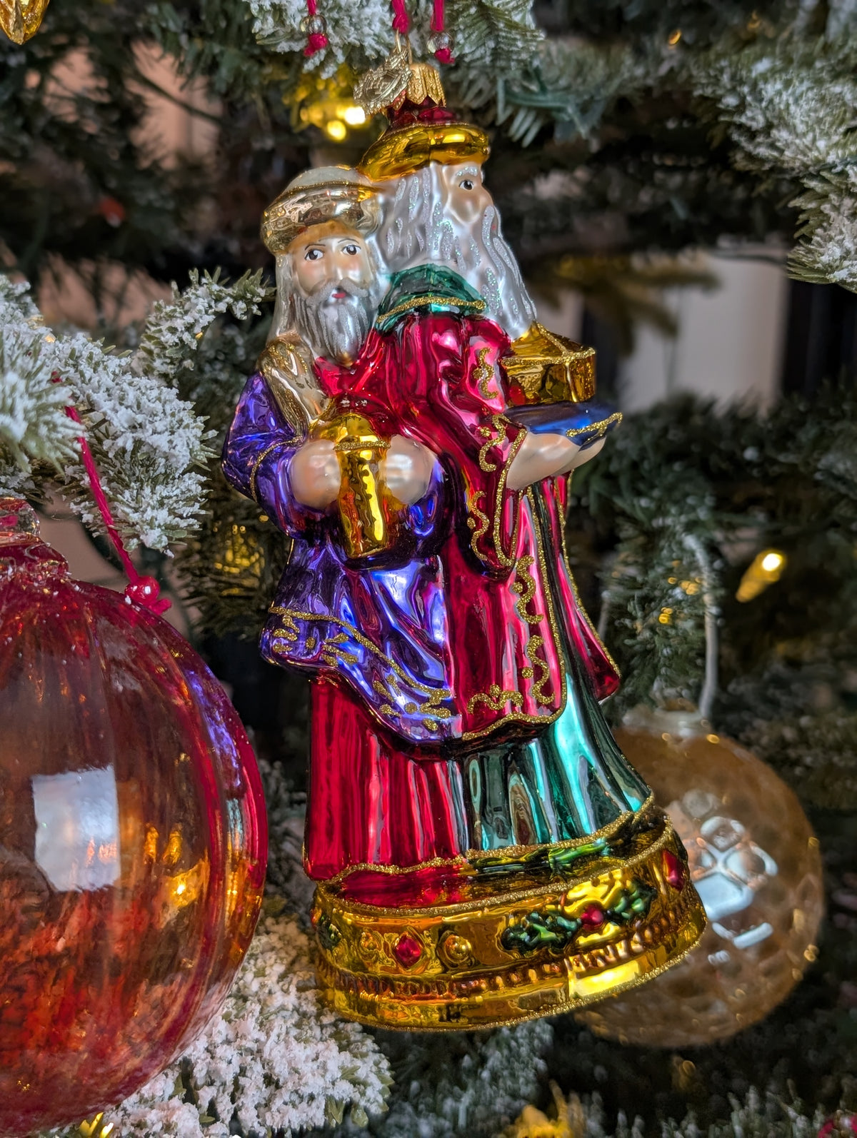 Suspension en forme des trois rois mages apportant leurs cadeaux. ils sont habillés de riches couleurs et chapeaux et sont posés sur un socle doré décoré de houx et baies rouges. Vue sur Gaspard de face apportant l'encens et Melchior de profil apportant l'or.