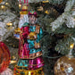 Suspension en forme des trois rois mages apportant leurs cadeaux. ils sont habillés de riches couleurs et chapeaux et sont posés sur un socle doré décoré de houx et baies rouges. Vue sur Balthazar de face apportant la myrrhe et Melchior de profil apportant l'or.