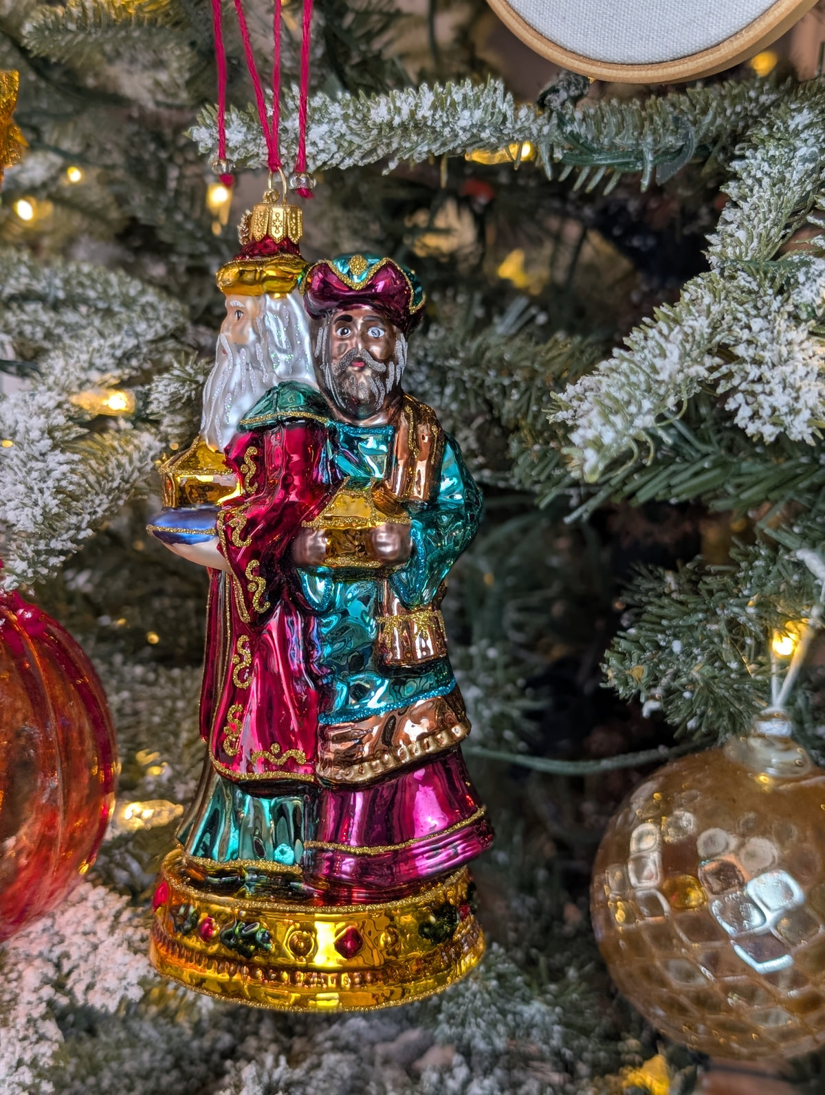 Suspension en forme des trois rois mages apportant leurs cadeaux. ils sont habillés de riches couleurs et chapeaux et sont posés sur un socle doré décoré de houx et baies rouges. Vue sur Balthazar de face apportant la myrrhe et Melchior de profil apportant l'or.