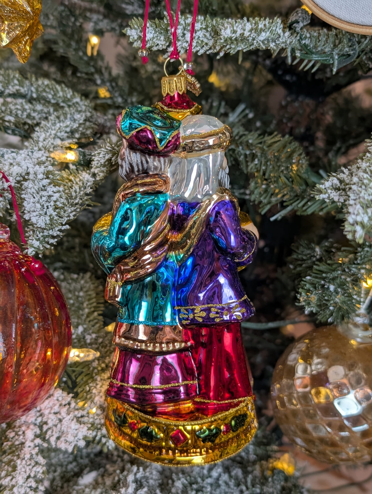 Suspension en forme des trois rois mages apportant leurs cadeaux. ils sont habillés de riches couleurs et chapeaux et sont posés sur un socle doré décoré de houx et baies rouges. Vue de Gaspard et Balthazar de dos.