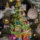 Suspension de Noël représentant un sapin de Noël décoré de guirlandes dorée en or 24 carats et de boules de Noël rouges, bleues, or et violettes et d'une étoile dorée au sommet. Des cadeaux bleus, violets et rouges, terminés par un nœud doré sont au pied du sapin. 