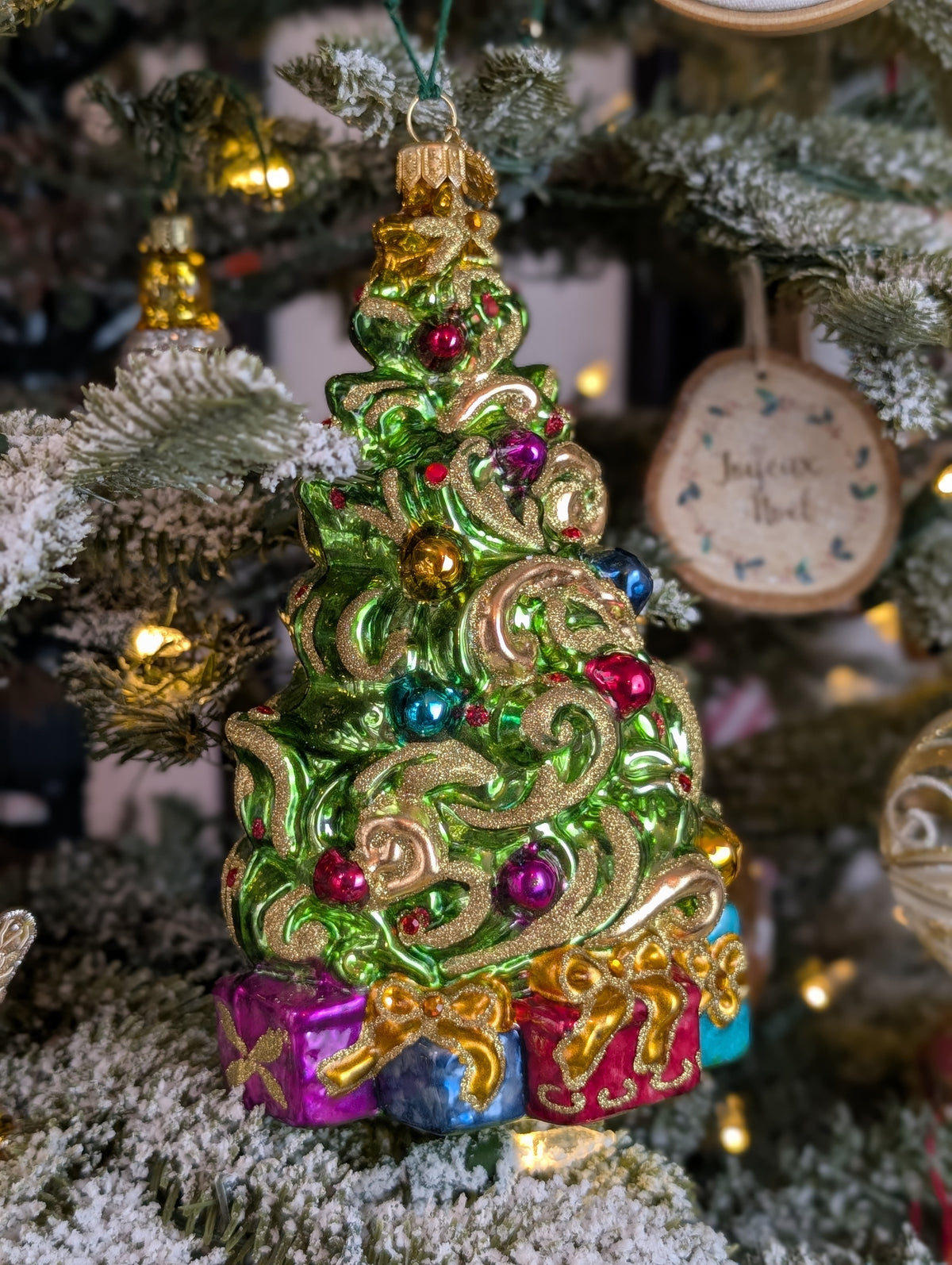 Suspension de Noël représentant un sapin de Noël décoré de guirlandes dorée en or 24 carats et de boules de Noël rouges, bleues, or et violettes et d'une étoile dorée au sommet. Des cadeaux bleus, violets et rouges, terminés par un nœud doré sont au pied du sapin. 