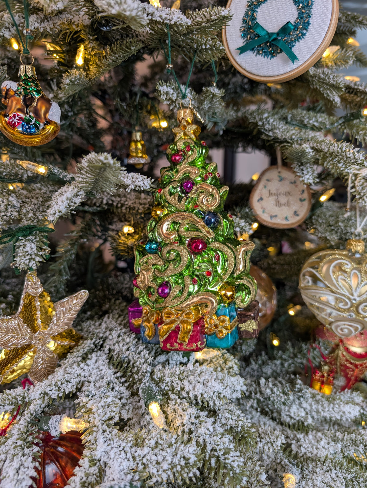 Suspension de Noël représentant un sapin de Noël décoré de guirlandes dorée en or 24 carats et de boules de Noël rouges, bleues, or et violettes et d'une étoile dorée au sommet. Des cadeaux bleus, violets et rouges, terminés par un nœud doré sont au pied du sapin.  Suspension en forme d'étoile en broderie d'or avec des paillettes de broderie dorées. Suspension brodée en forme de couronne de noël verte avec un nœud et des perles verts et de la canetille or. Une suspension miniature en  forme de cheval.