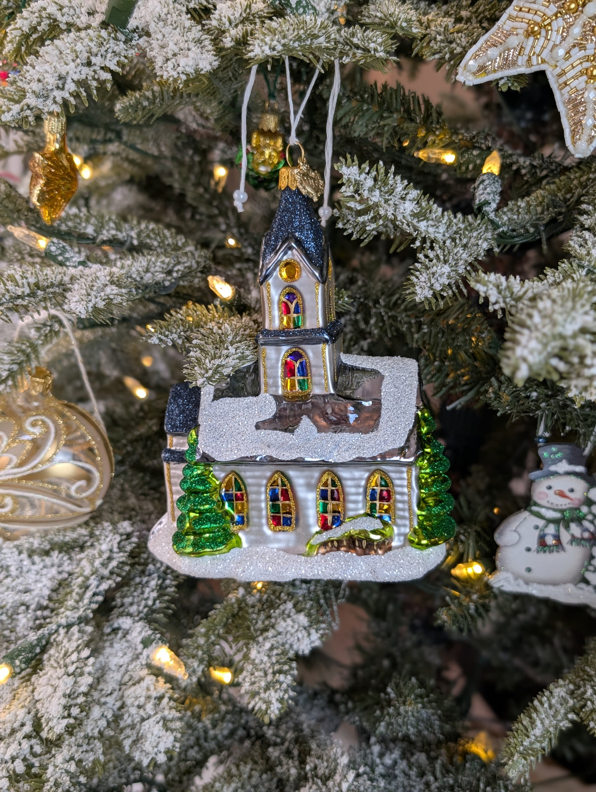 Suspension en forme d'église blanche, avec des vitraux colorés vert, jaune, bleu, violet et rouge et des rosaces dorées.  le toit bleu gris est enneigé et l'église est entourée de sapin verts