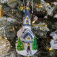 Suspension en forme d'église blanche, avec des vitraux colorés vert, jaune, bleu, violet et rouge et des rosaces dorées.  le toit bleu gris est enneigé et l'église est entourée de sapin verts. 