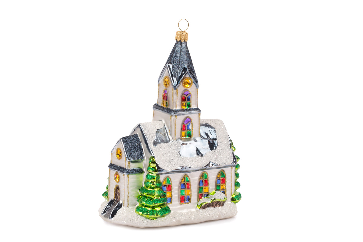 Suspension en forme d'église blanche, avec des vitraux colorés vert, jaune, bleu, violet et rouge et des rosaces dorées.  le toit bleu gris est enneigé et l'église est entourée de sapin verts