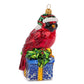 Suspension en forme d'oiseau cardinal rouge qui porte un bonnet de noël vert rayé de rouge avec fourrure et pompon blanc, assis sur un cadeau bleu décoré de flocons blancs et sertie d'un nœud doré et de feuilles de houx.
