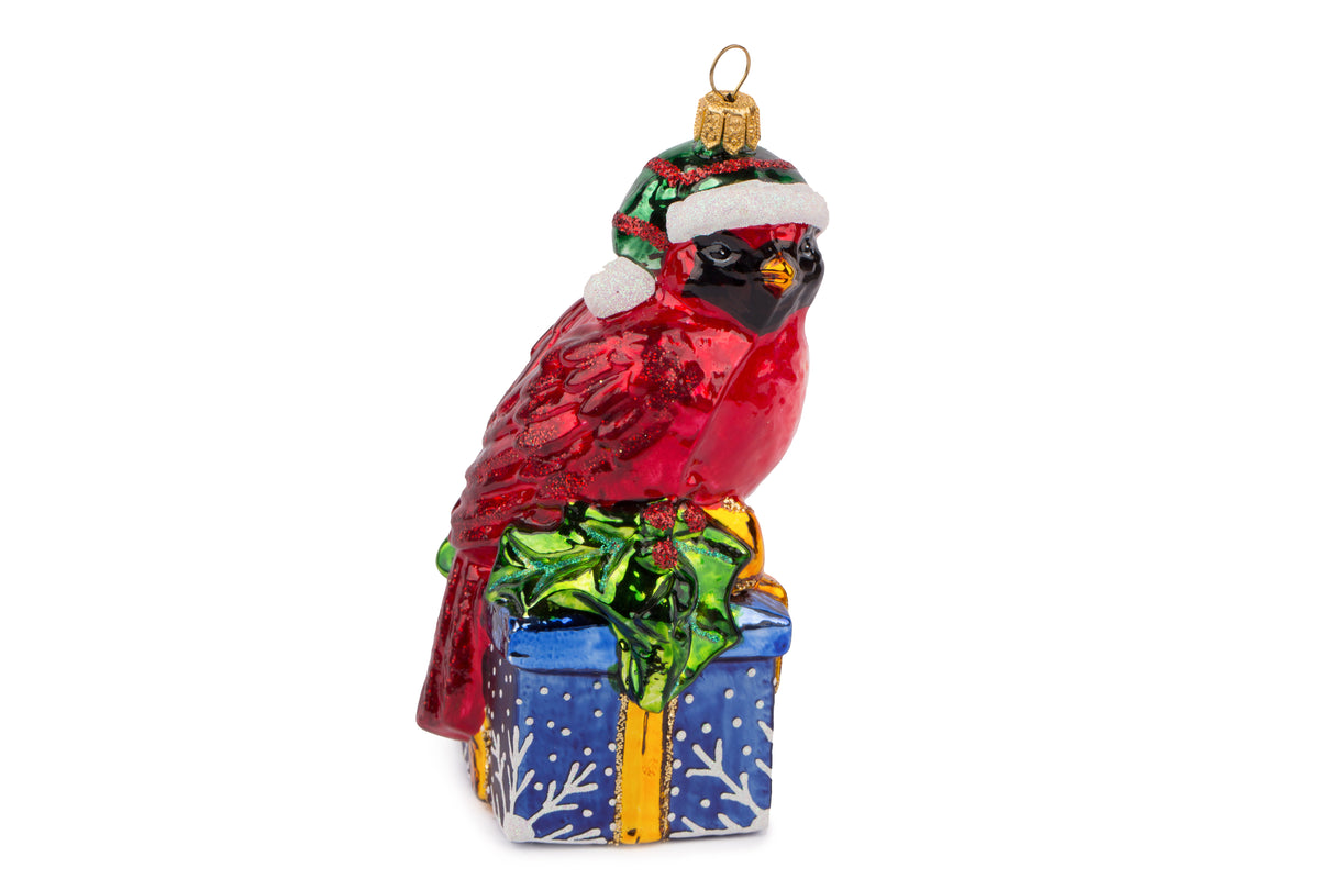 Suspension en forme d'oiseau cardinal rouge qui porte un bonnet de noël vert rayé de rouge avec fourrure et pompon blanc, assis sur un cadeau bleu décoré de flocons blancs et sertie d'un nœud doré et de feuilles de houx.