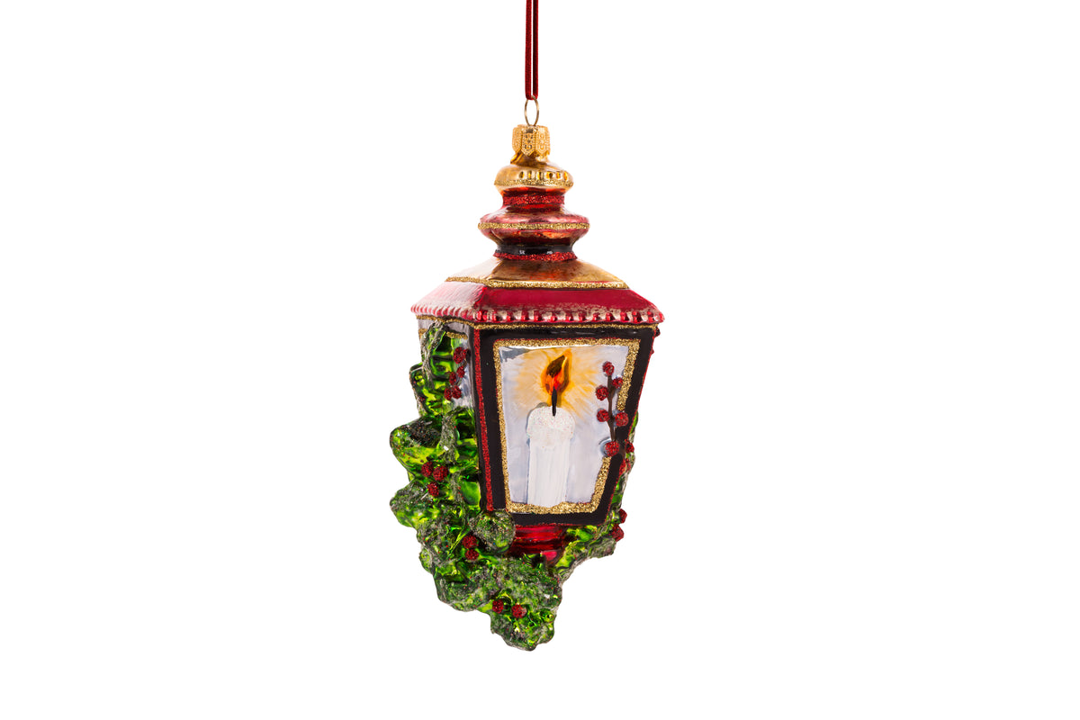 Suspension en forme de lanterne rouge et or avec une bougie allumée à l'intérieur et des branches de sapin et baies rouges sur les cotés. 